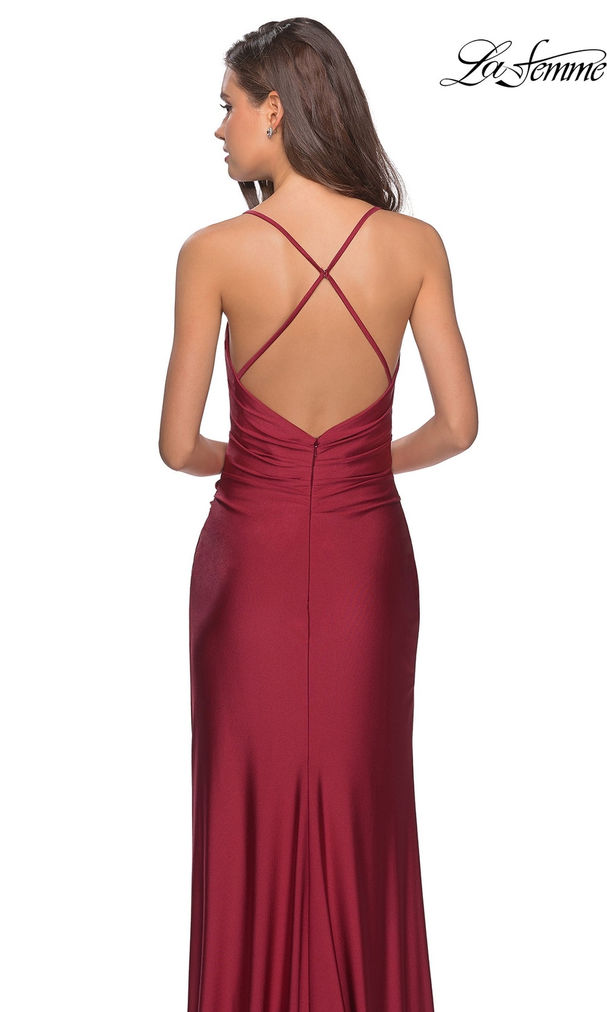A woman stands with her back to the camera in the La Femme Open-Back Long V-Neck Prom Dress 28206, a sleeveless and backless burgundy gown featuring crisscross straps, a fitted silhouette, slight ruching, and elegant prom style.